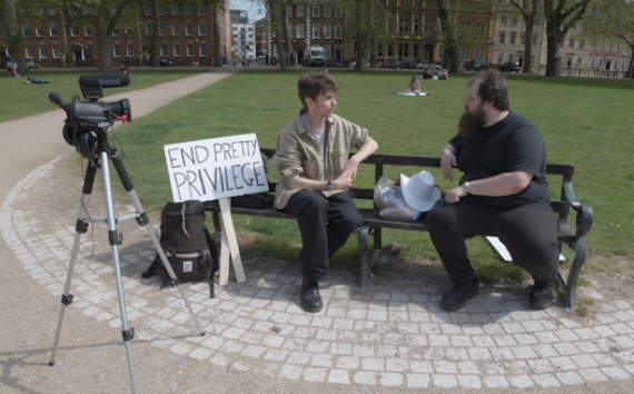 Interview being conducted during the Ugly Rights March with 'END PRETTY PRIVILEGE' sign visible